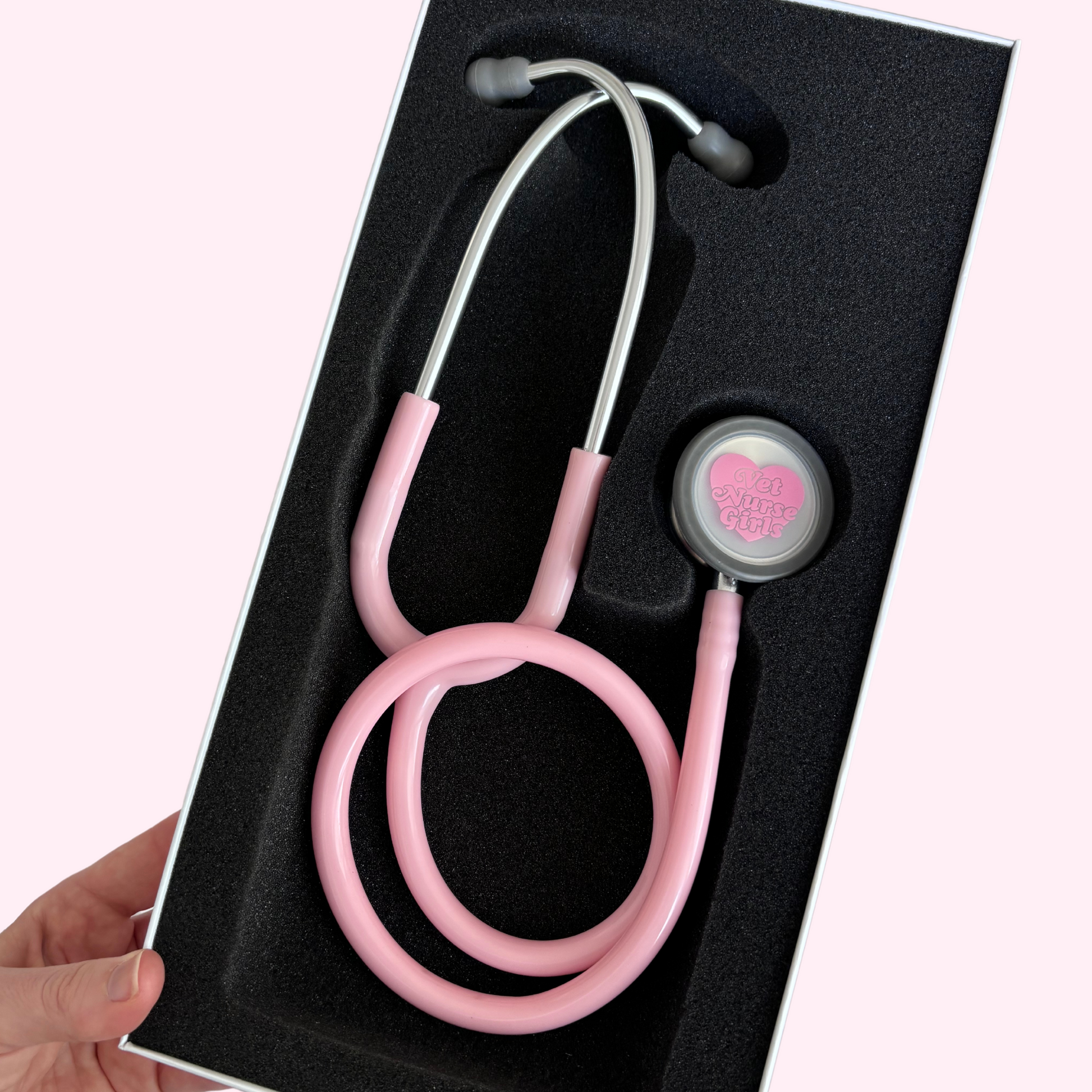 A baby pink stethoscope with a dual-head design, presented in a black box with a clear plastic window showing the instrument. It has stainless steel tubing and PVC ear tips.